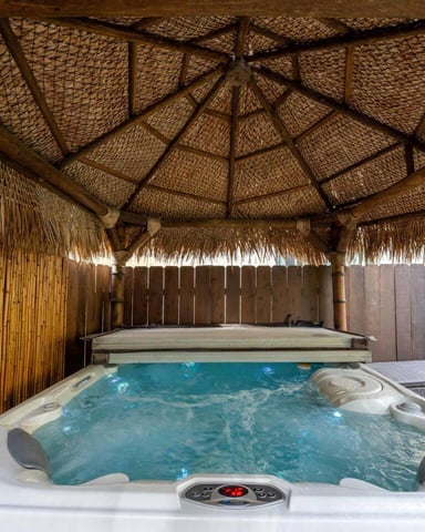 Hot tub with jets under thatched roof shelter, wooden fence and tropical plants visible
