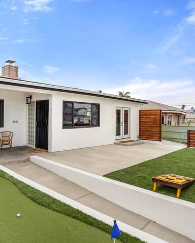 Modern single-story white house with concrete patio, artificial turf yard, cornhole game, and wooden fence