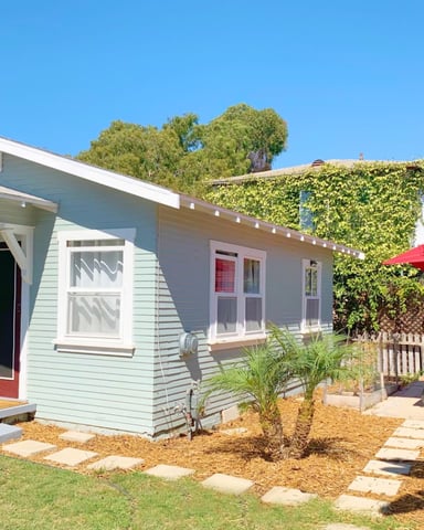 Light blue cottage with red door and porch, backyard with red umbrella, grill, and ivy-covered fence