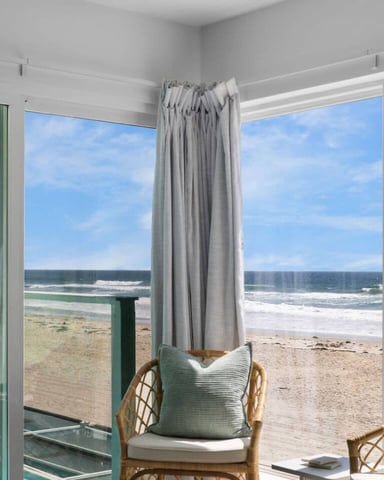 Bright room with floor-to-ceiling windows overlooking sandy beach, ocean waves, and blue sky, with wicker chairs and gray curtains
