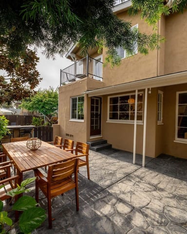 Tan stucco house with patio featuring wooden dining table, chairs, and mature trees overhead