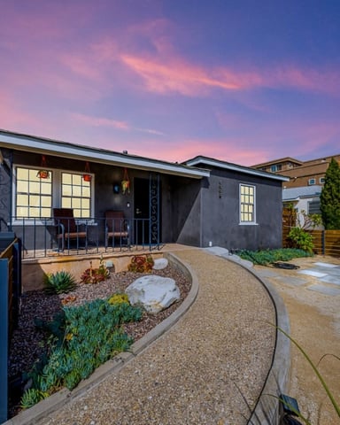 Modern dark gray house with illuminated windows at dusk, featuring a front patio with desert landscaping and wooden fencing.