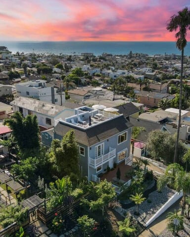 Aerial view of coastal neighborhood with white house, palm trees, and ocean sunset in background