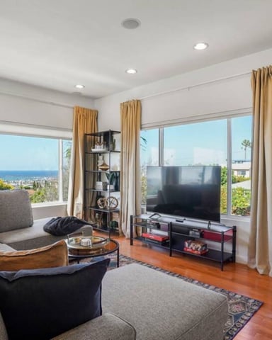 Modern living room with gray sectional sofa, TV, black shelving, hardwood floors, and ocean views through large windows with gold curtains