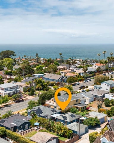 Aerial view of coastal neighborhood with single-family homes, palm trees, and ocean in background, yellow location pin marking property