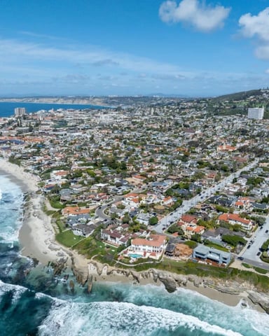Aerial view of coastal suburb with sandy beach, rocky cliffs, residential homes, and ocean waves under blue sky