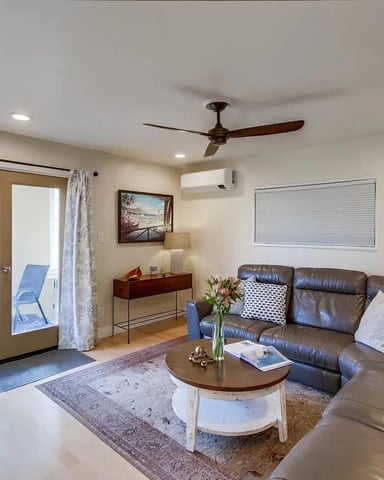 Bright living room with brown leather sectional sofa, round coffee table, glass doors opening to balcony with water view, ceiling fan, and wall clock.