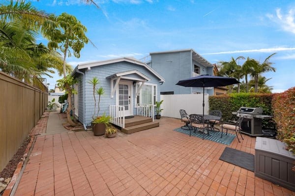 Light blue cottage with white trim and deck, surrounded by palm trees and tropical plants, brick patio with outdoor seating and grill