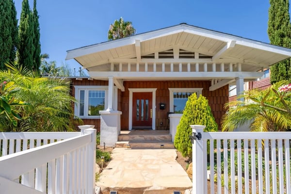 Modern mid-century home with brown wood siding, white trim, and overhanging eaves, flanked by white picket fencing and tropical landscaping
