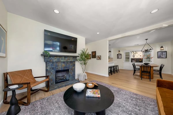 Open-plan living room with blue tile fireplace, wall-mounted TV, leather chair, round black coffee table, and dining area with wood table visible in background.
