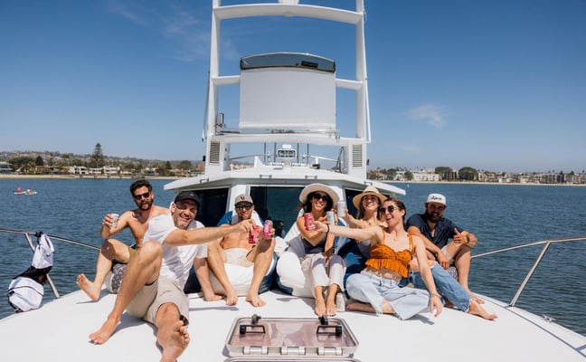 Group of seven people sitting on the deck of a white fishing boat in calm blue water with coastal homes visible in the background.